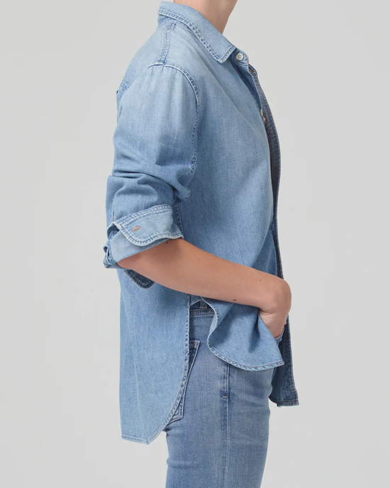 Side view of a person wearing a denim Shrunken Kayla Shirt in Tide by Citizens of Humanity with rolled-up sleeves and hands in pockets, featuring an oversized fit.