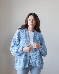 Woman in a blue handknit Placid cardigan by Margaret O'Leary standing against a neutral background.