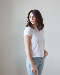 A woman in a Velvet by Graham & Spencer Sierra S/S Crew Neck Top in White and blue jeans standing against a neutral background, looking to the side.
