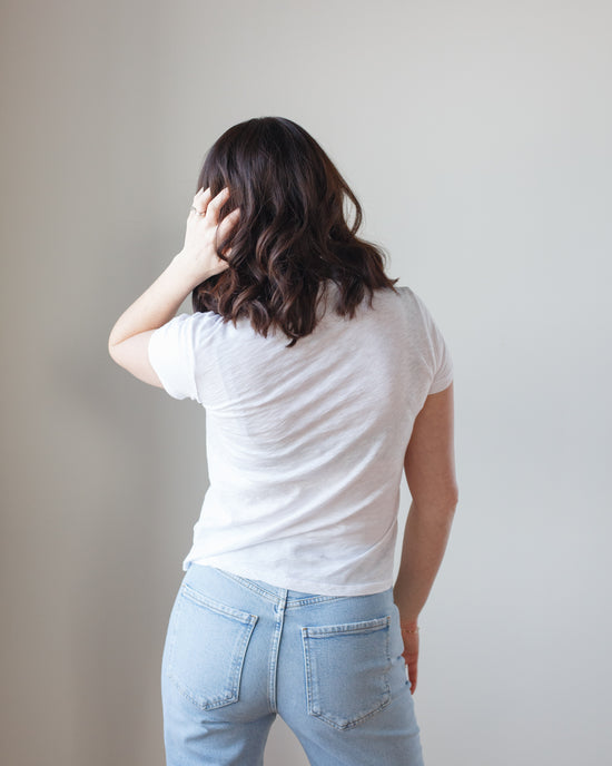 Woman in a Velvet by Graham & Spencer Sierra S/S Crew Neck Top in White and blue jeans standing with her back to the camera and hand on her head.