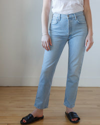 A person wearing AGOLDE Riley Long in Issue high rise straight light blue jeans, a white top, and black open-toe sandals standing in front of a neutral background.