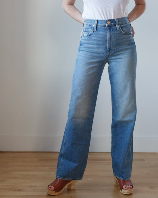 Person wearing AMO Tricia Wide Straight in Joyous blue jeans and brown sandals standing against a white wall.