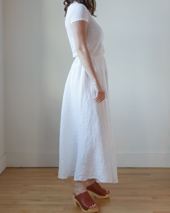 Woman standing sideways wearing a CP Shades Deidra Skirt HW Linen Twill in White midi dress and brown platform sandals.