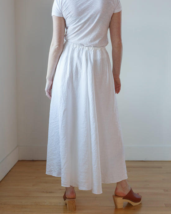 Woman standing in a CP Shades Deidra Skirt HW Linen Twill in White and clogs, facing away from the camera.