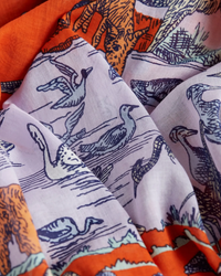 Close-up of a 100% Cotton fabric with bird patterns in purple on a white background, partially covered by an Inoui Editions' Scarf 100 Reverie in Terracotta with contrasting designs.
