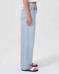 A person wearing light blue denim AGOLDE Low Slung Baggy jeans and black sandals with white straps stands sideways.