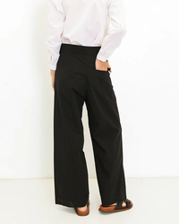 A person standing with their back facing the camera, wearing Marnie - Parachute in Black trousers, a white shirt, and brown sandals by A Shirt Thing.