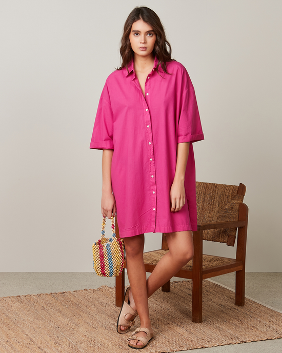 A woman stands next to a wooden chair, modeling an oversized short sleeve Hartford Rimo Dress in Hibiscus with sandals and holding a colorful woven bag.