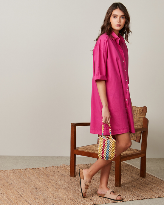 Woman in a Hartford Rimo Dress in Hibiscus with oversized short sleeves holding a multicolored bag standing beside a wooden chair.