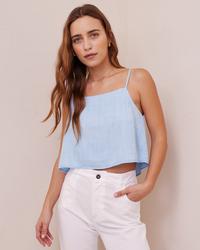 A woman wearing a blue Button Back Cami in Paradis Wash by Bella Dahl with a square neckline and white pants against a neutral background.