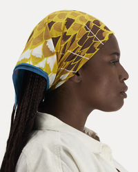 A woman in profile wearing an Inoui Editions Square 65 Neofelis bandana in Yellow as a patterned headscarf and a white shirt.