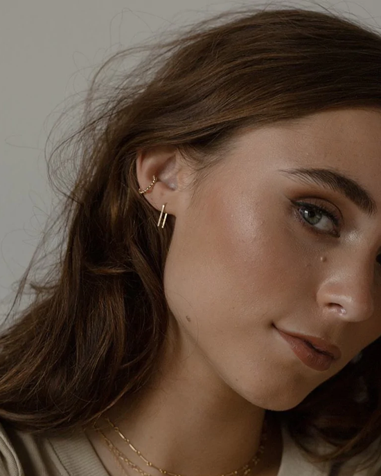 Close-up of a woman with subtle makeup and a Token Jewelry Mini Staple Earrings in 14K Gold Fill, featuring a side view of her face with brown hair tucking behind her ear.