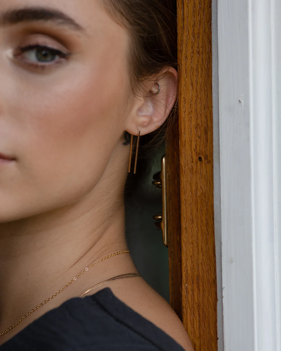 Close-up of a woman's face partially obscured by a door frame, highlighting her Staple Earrings in 14K Gold Fill by Token Jewelry.
