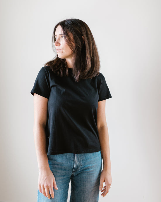 A woman in a Frankie Tee in Black by Velvet by Graham & Spencer and blue high rise straight leg jeans standing against a neutral background, looking to her right.