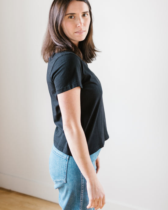 A woman in a Velvet by Graham & Spencer Frankie Tee in Black and high rise straight leg jeans standing against a white wall, looking over her shoulder at the camera.