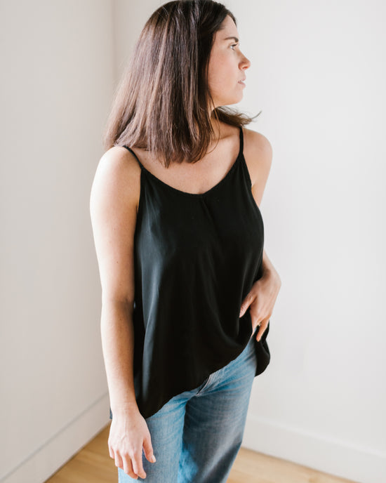 A woman in a black CP Shades Fairie Tank with a high-low hem and blue jeans standing inside and looking to the side.