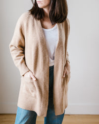 Woman wearing a beige 100% Cashmere Margaret O'Leary Luxe Sweater Coat, white top, and blue jeans standing against a neutral background.