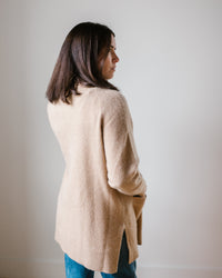 A woman in a Margaret O'Leary Luxe Sweater Coat in Camel and blue jeans standing sideways against a neutral background.
