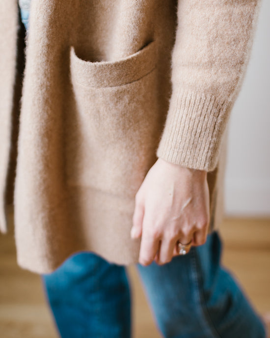 A person standing with a hand in the pocket of a luxe Luxe Sweater Coat in Camel by Margaret O'Leary.