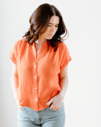 Woman standing against a white background, wearing a Felicite Apparel Cherry Short Sleeve Top and blue jeans, looking down to her side.