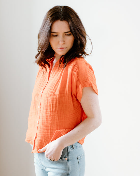 A woman wearing a Felicite Apparel Short Sleeve Top in Cherry with a Mandarin collar and blue jeans is standing against a neutral background, looking to her right with a contemplative expression.