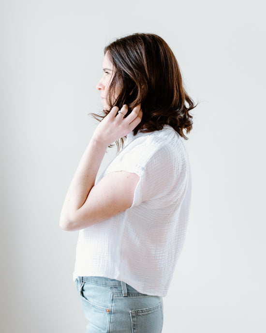 A woman in a Felicite Apparel short sleeve top in white and blue jeans standing sideways, touching her neck against a plain background.