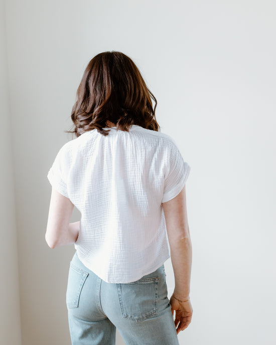 Woman standing with her back to the camera, wearing a Felicite Apparel Short Sleeve Top in White against a white background.