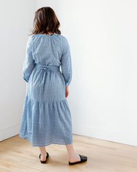 Woman standing facing a white wall, wearing a Felicite Apparel Puff Sleeve Maxi Dress in Sky with a detachable waist tie and black flats.