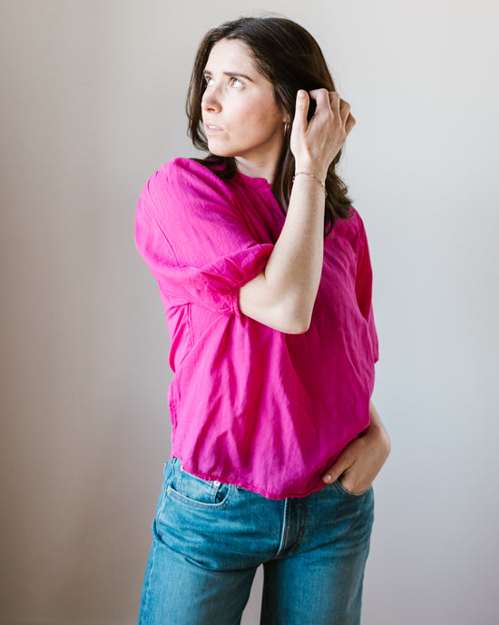 A woman in a bright pink Hartford Hibiscus Shirt with a mandarin collar and blue jeans stands against a neutral background, looking to the side with her hand in her hair.