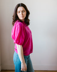 A woman in a pink Hartford Turette Shirt in Hibiscus with a mandarin collar and blue jeans standing with her side toward the camera, her eyes closed and head slightly tilted downwards.