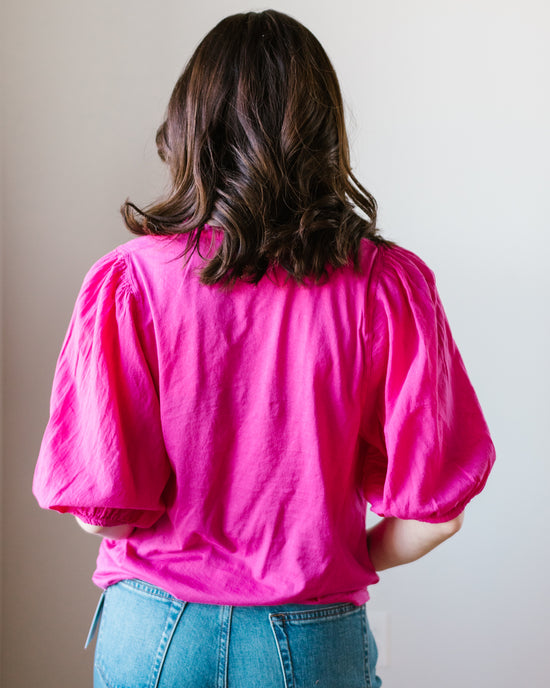 Woman standing with her back to the camera, wearing a bright pink Hartford Turette Shirt in Hibiscus with puffy sleeves and blue jeans, perfect for a business casual look.