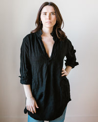 A woman standing against a neutral background, wearing a sustainable CP Shades Teton in Black Heavy Weight Linen Twill blouse and denim skirt, with her hair down.