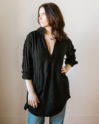 A woman in a loose-fitting black CP Shades Teton Tunic and blue jeans standing against a neutral background.