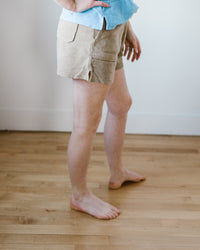 A person standing in Hartford Timoe Shorts in Dune and a blue shirt with their hands in their pockets.