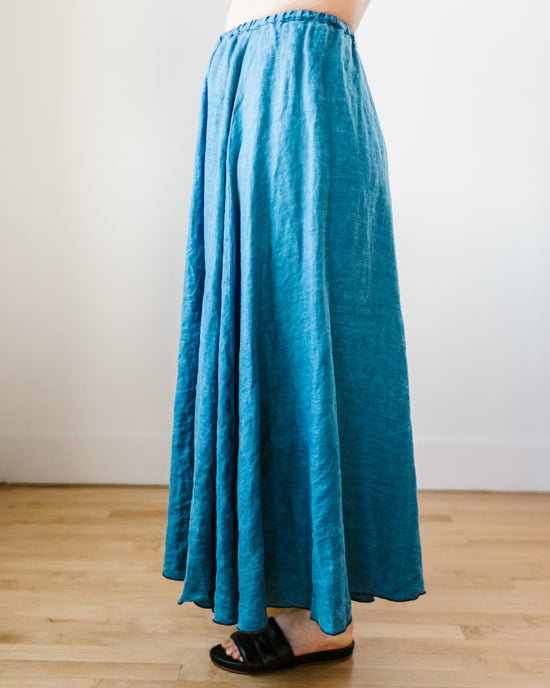 A person standing in a room wearing a long blue Deidra Skirt in Bleach Indigo Twill by CP Shades and black sandals.