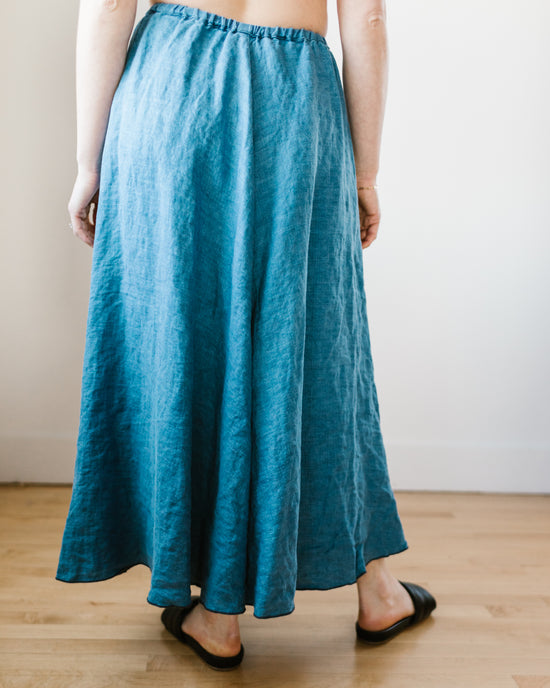 A person standing in a room wearing a long blue CP Shades Deidra Skirt in Bleach Indigo Twill and black flat shoes.