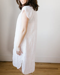 A woman in a white CP Shades Lucy Dress w/o Pkts in White HW Linen Twill standing sideways with a neutral background.