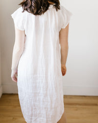 A person standing facing away from the camera, wearing a white, knee-length 100% Linen Lucy Dress w/o Pkts in White HW Linen Twill by CP Shades with short sleeves, against a plain background.