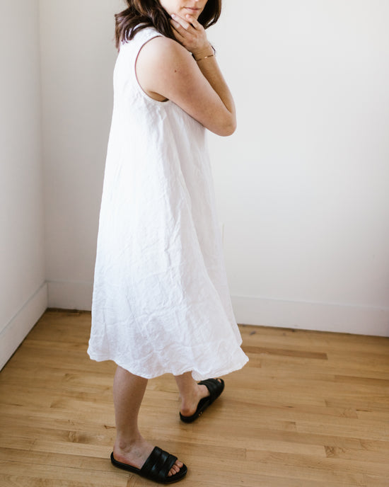 Woman in a sleeveless Bree Dress in White HW Linen Twill by CP Shades standing in a room with wooden flooring.