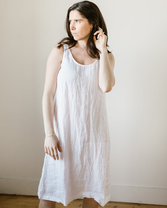 A woman in a white CP Shades Dayna Dress in White HW Linen Twill standing beside a plain wall, looking to the side with her hand near her face.