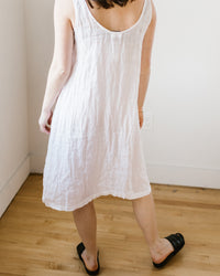 Woman wearing a CP Shades Dayna Dress in White HW Linen Twill and black slides standing in a room.