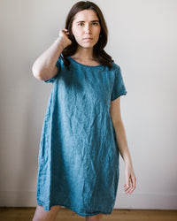 A woman in a blue CP Shades Esme Dress w/o Pockets in Bleach Indigo Twill standing against a plain background.