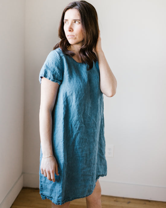 A woman in a blue CP Shades Esme Dress w/o Pockets in Bleach Indigo Twill standing against a white wall, looking to the side.