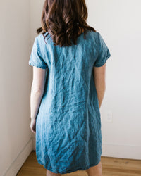 Woman standing indoors wearing a CP Shades Esme Dress w/o Pockets in Bleach Indigo Twill, facing away from the camera.