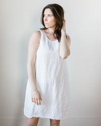 Woman in a Jess Dress in White HW Linen Twill by CP Shades standing against a neutral background, looking to the side with her hand in her hair.