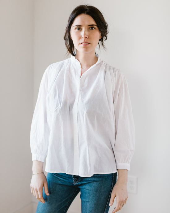 Woman in a casual Flora - Cabo in White blouse by A Shirt Thing and blue jeans standing against a white wall.