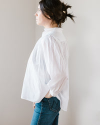 Woman standing sideways in a Flora - Cabo in White 100% cotton, relaxed fit blouse from A Shirt Thing and jeans against a neutral background.