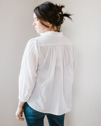 Woman seen from behind, wearing a Flora - Cabo in White button-down blouse and blue jeans, with hair in a ponytail.
