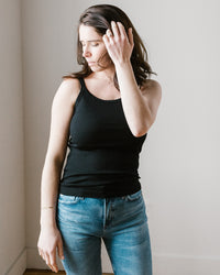 A woman in a black Aliza Tank in Black by Velvet by Graham & Spencer and blue jeans standing beside a wall with her hand in her hair.