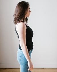 A woman in a black Cotton Slub Aliza Tank Top by Velvet by Graham & Spencer and blue jeans standing side-on to the camera against a neutral background.